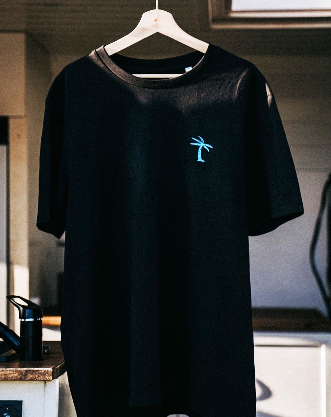 Strandiesdas Palma One T-Shirt in Schwarz mit türkiser Palme hängt am Fenster